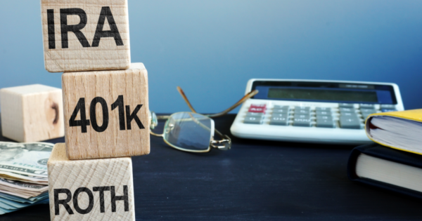 A desk with a calculator, glasses, money and 3 blocks with IRA, 401k and ROTH written on them. These are meant to represent a guide to the many different kinds of retirement plans.